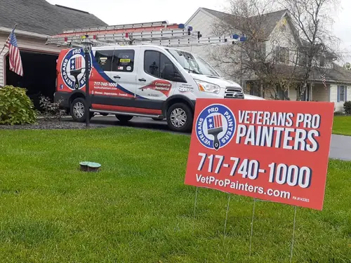 Veterans Pro Painters van with ladders on the roof rack in a customer's driveway, red yard sign on the lawn